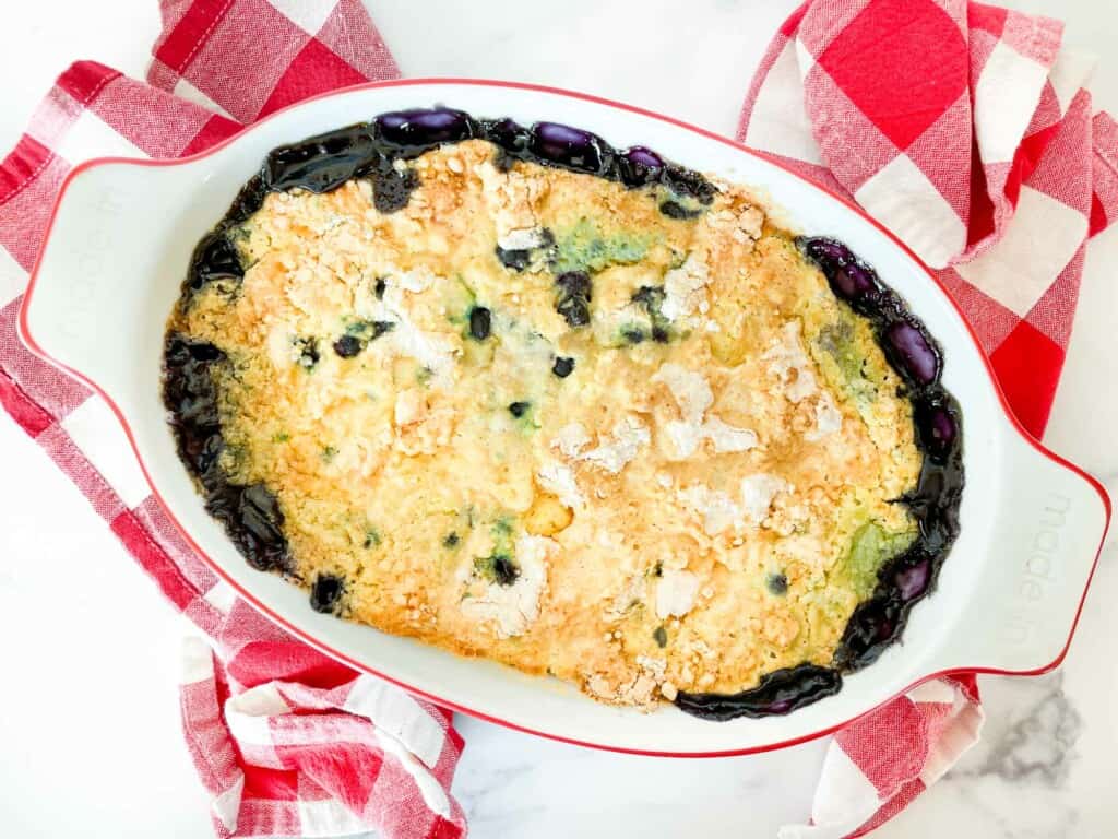 A baked blueberry cobbler with a golden, crumbly topping in a white oval dish, surrounded by red and white checkered towels.