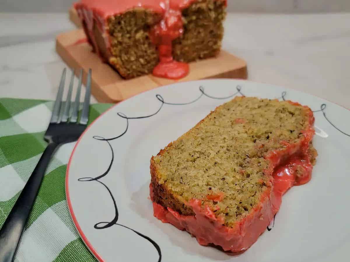 A slice of banana bread with pink icing sits on a plate in the foreground, with the rest of the loaf and a fork nearby.