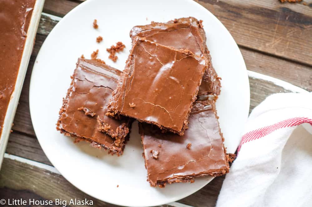 Three chocolate frosted brownies stacked on a white plate, next to a white cloth with red stripes on a wooden surface.