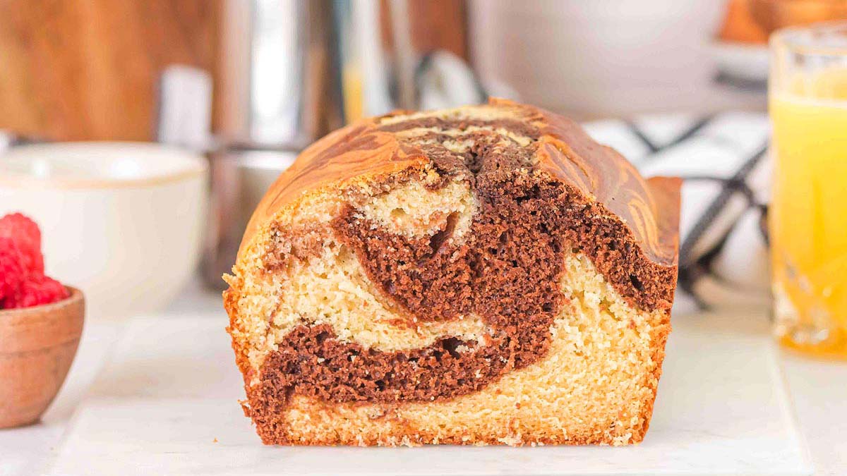 A close-up of a sliced marble loaf cake showing swirls of chocolate and vanilla on a light countertop, with a bowl of raspberries and a glass of orange juice in the background.