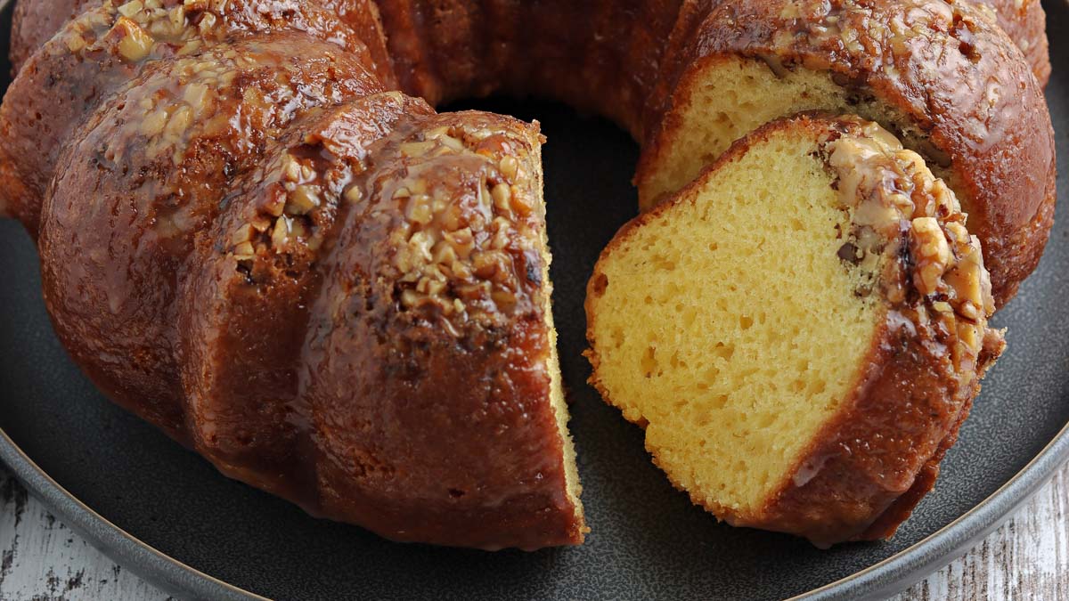 A bundt cake with a slice cut out, showing a moist yellow interior and a glazed topping with chopped nuts, on a dark plate.