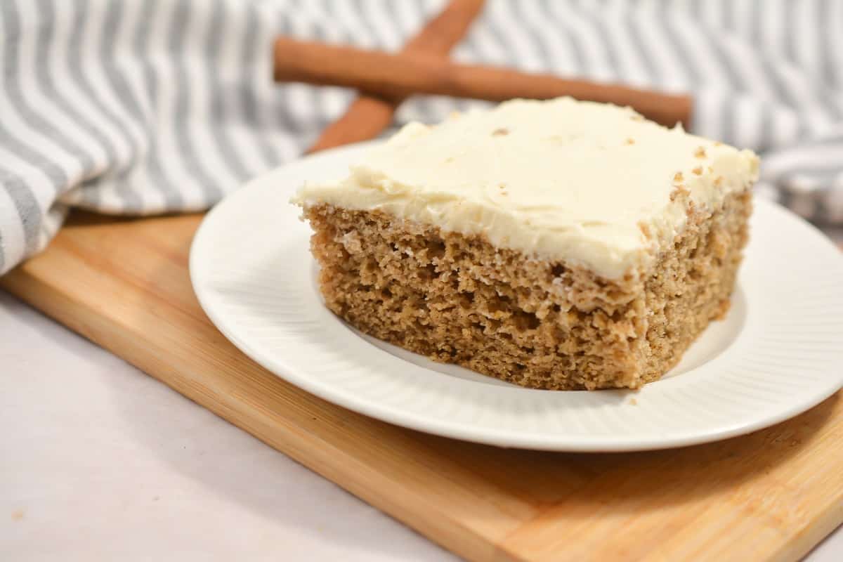 A square slice of frosted cake sits on a white plate atop a wooden board, with a striped cloth and two cinnamon sticks in the background.