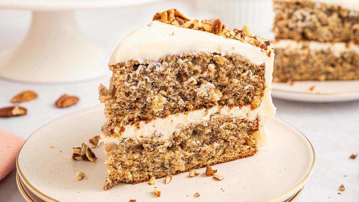A slice of two-layer cake with white frosting and chopped nuts on top, served on a white plate.