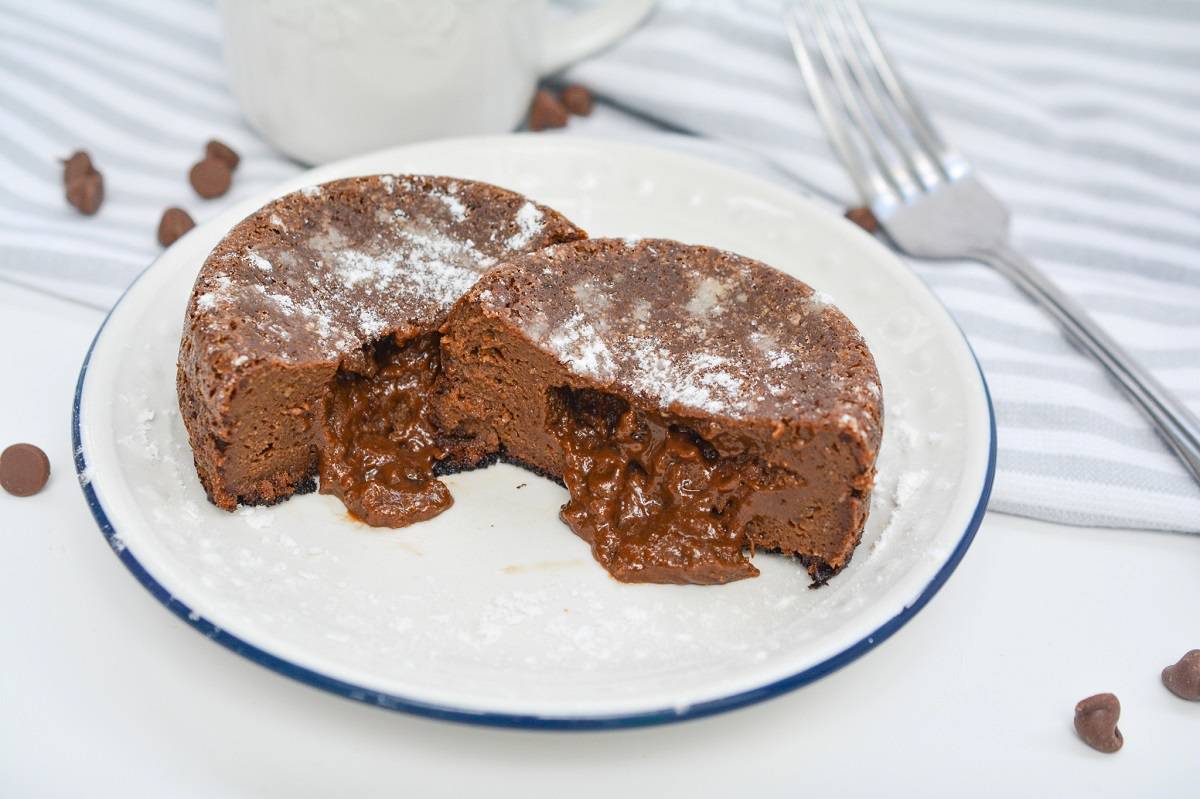 A chocolate lava cake cut in half on a white plate, with powdered sugar on top and melted chocolate filling visible inside. Chocolate chips and utensils are in the background.