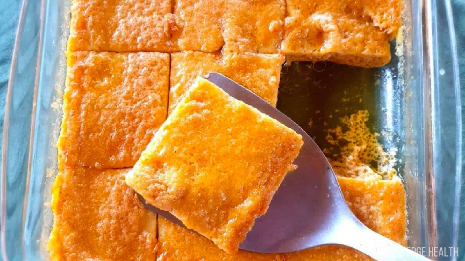 A glass baking dish contains several square pieces of baked cornbread, with one piece being lifted by a metal spatula.