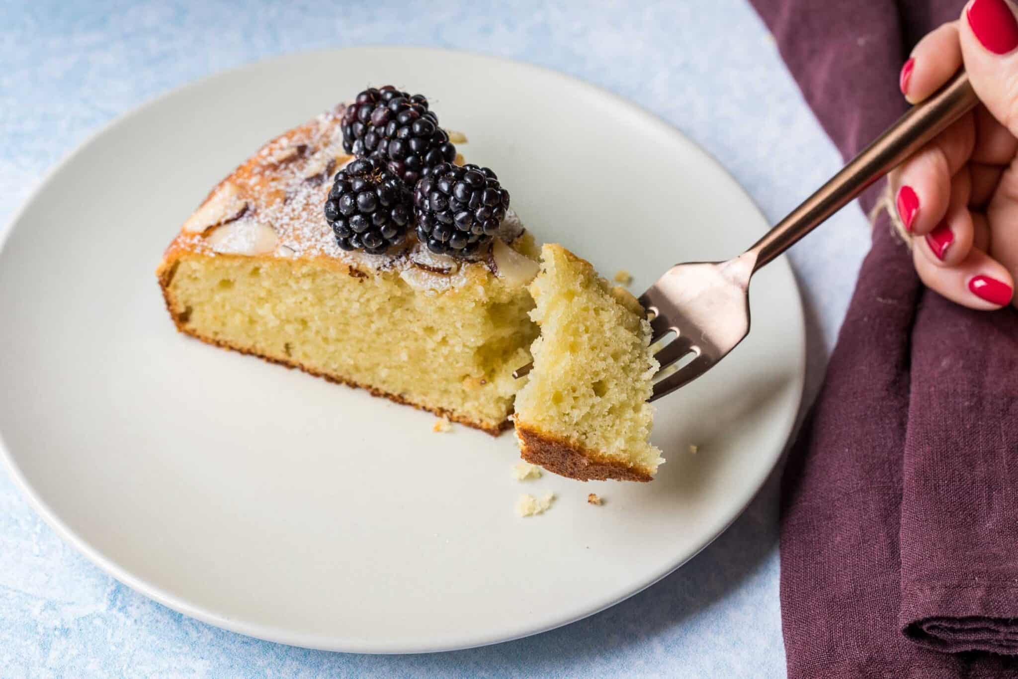 A slice of sponge cake topped with powdered sugar and blackberries on a white plate, with a fork holding a bite-sized piece.