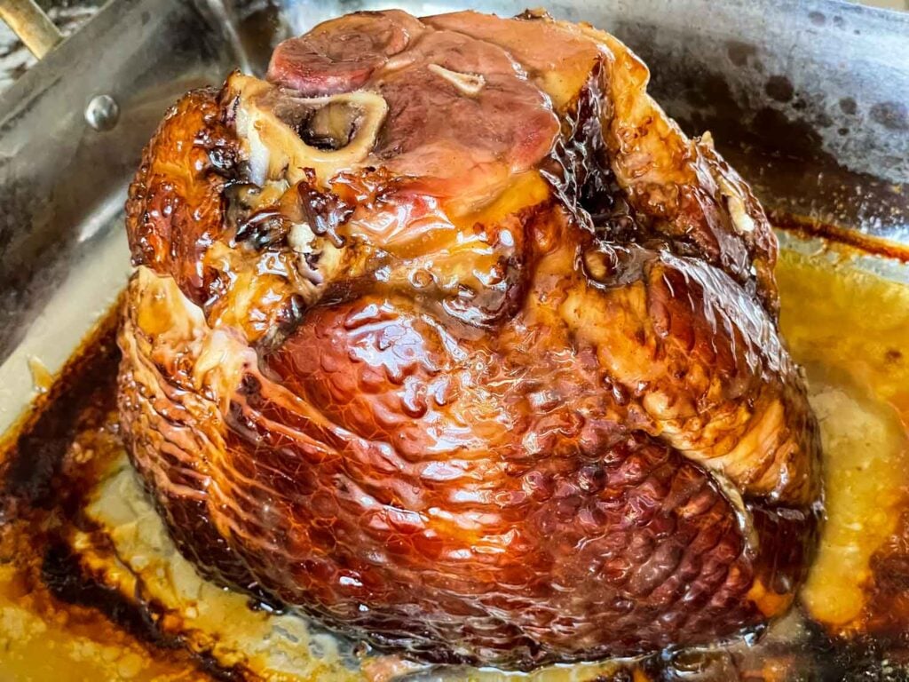 A glazed, baked ham with a visible bone sits in a metal roasting pan, surrounded by juices and caramelized edges.