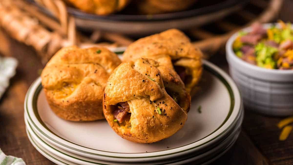 Three baked muffin-shaped pastries with a golden crust and savory filling are arranged on a stack of plates, with a bowl of diced ingredients in the background.