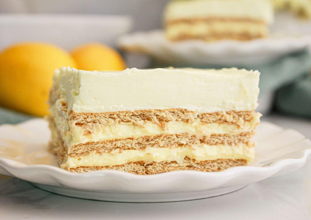 A slice of layered lemon icebox cake with graham crackers and creamy filling is served on a white plate; whole lemons are visible in the background.