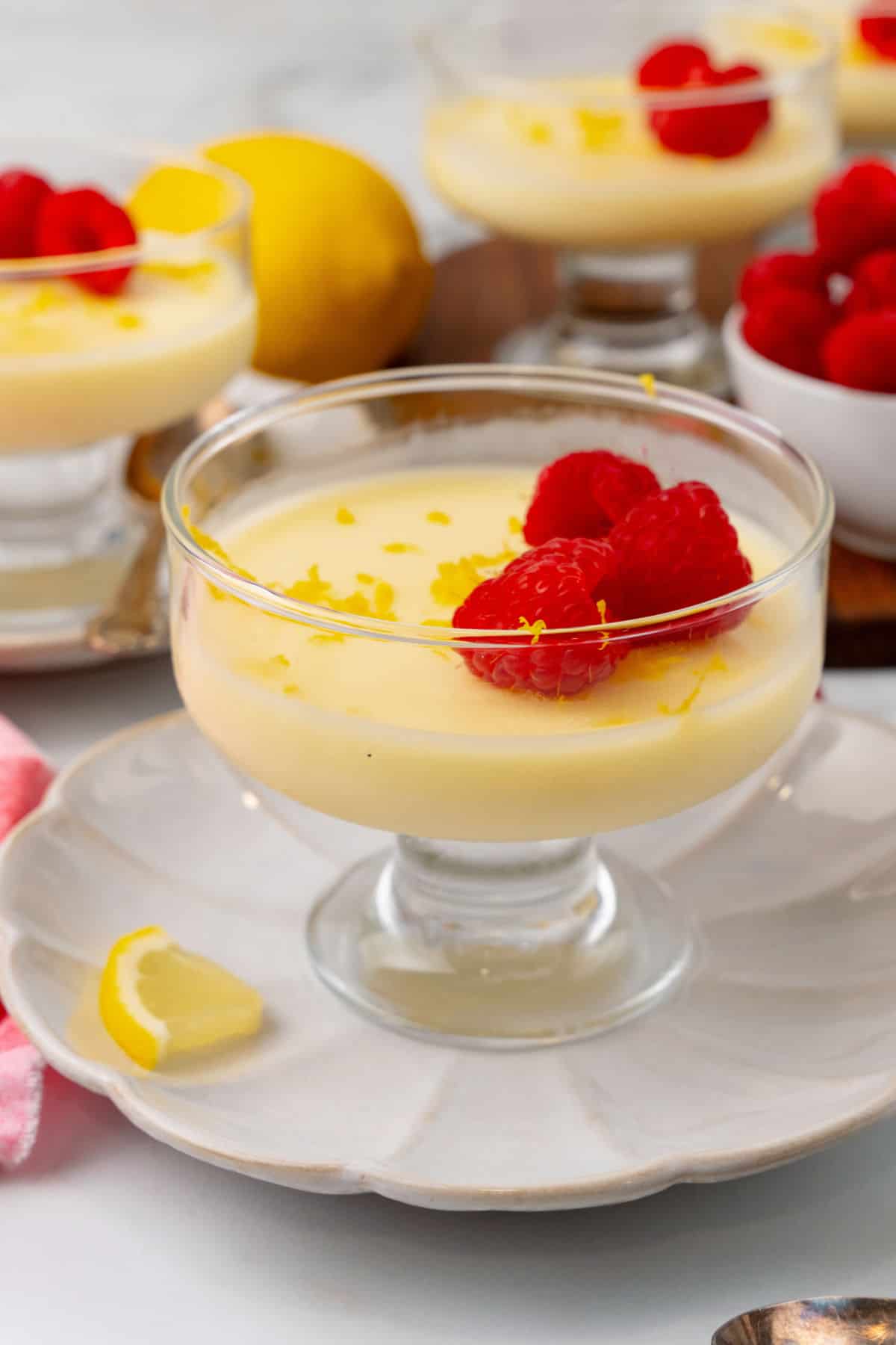 Glass dessert dish with lemon pudding, topped with fresh raspberries and lemon zest, placed on a white plate with a lemon wedge and additional desserts in the background.