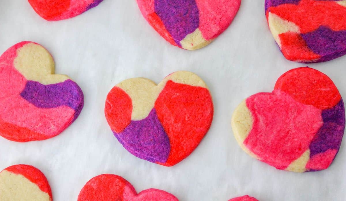 Heart-shaped cookies with a marbled pattern in red, pink, purple, and white are arranged on a white surface.