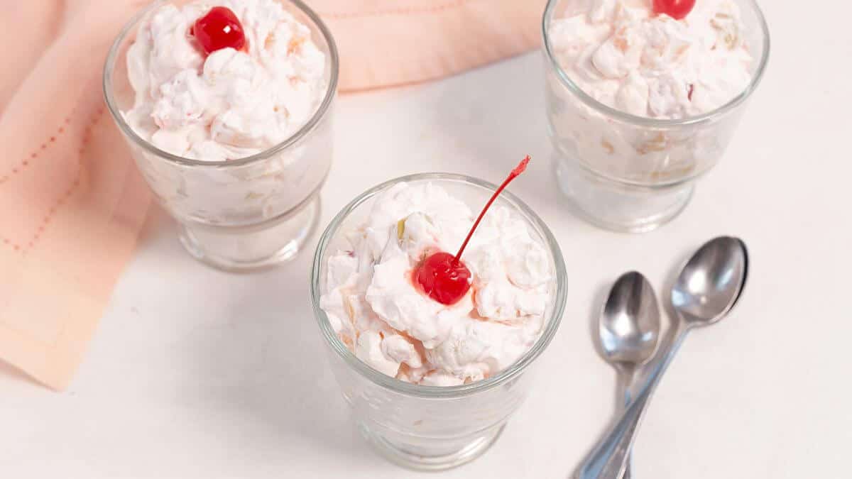 Three glass cups filled with whipped fruit salad, each topped with a maraschino cherry, next to two metal spoons on a white surface.