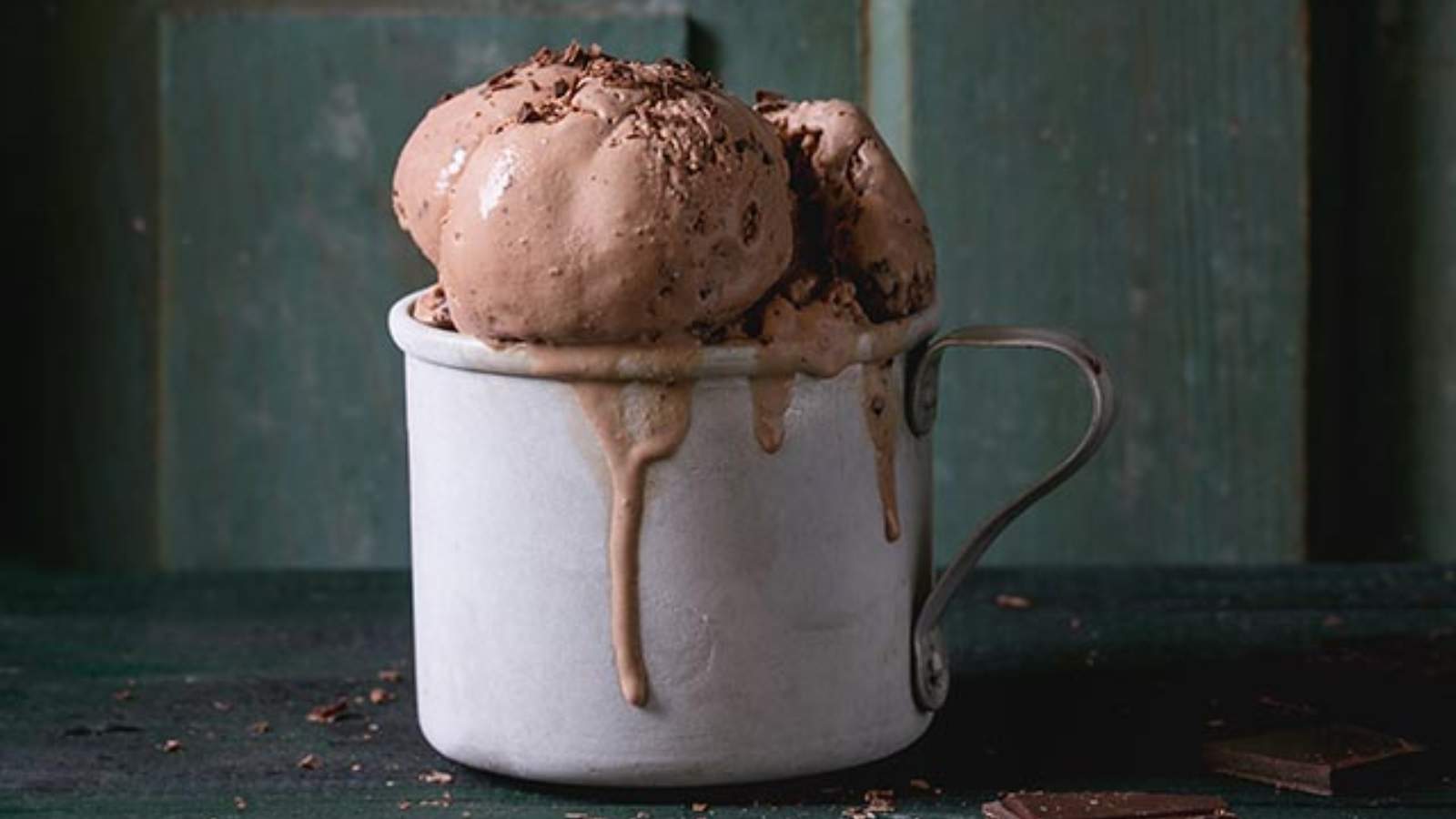 A metal cup filled with scoops of chocolate ice cream, with some melted ice cream dripping down the side, set against a dark green wooden background.