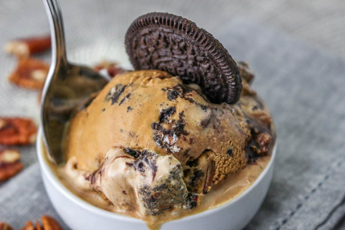 A bowl of ice cream with chocolate cookie chunks, a whole chocolate sandwich cookie on top, and a spoon, set on a gray surface with pecans in the background.