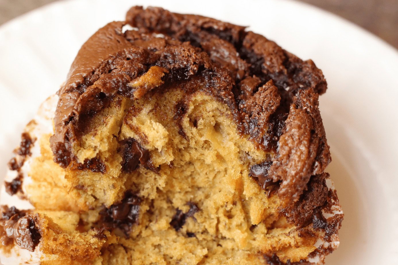 Close-up of a chocolate chip muffin with a bite taken out, showing a moist, crumbly texture and swirls of chocolate on top.