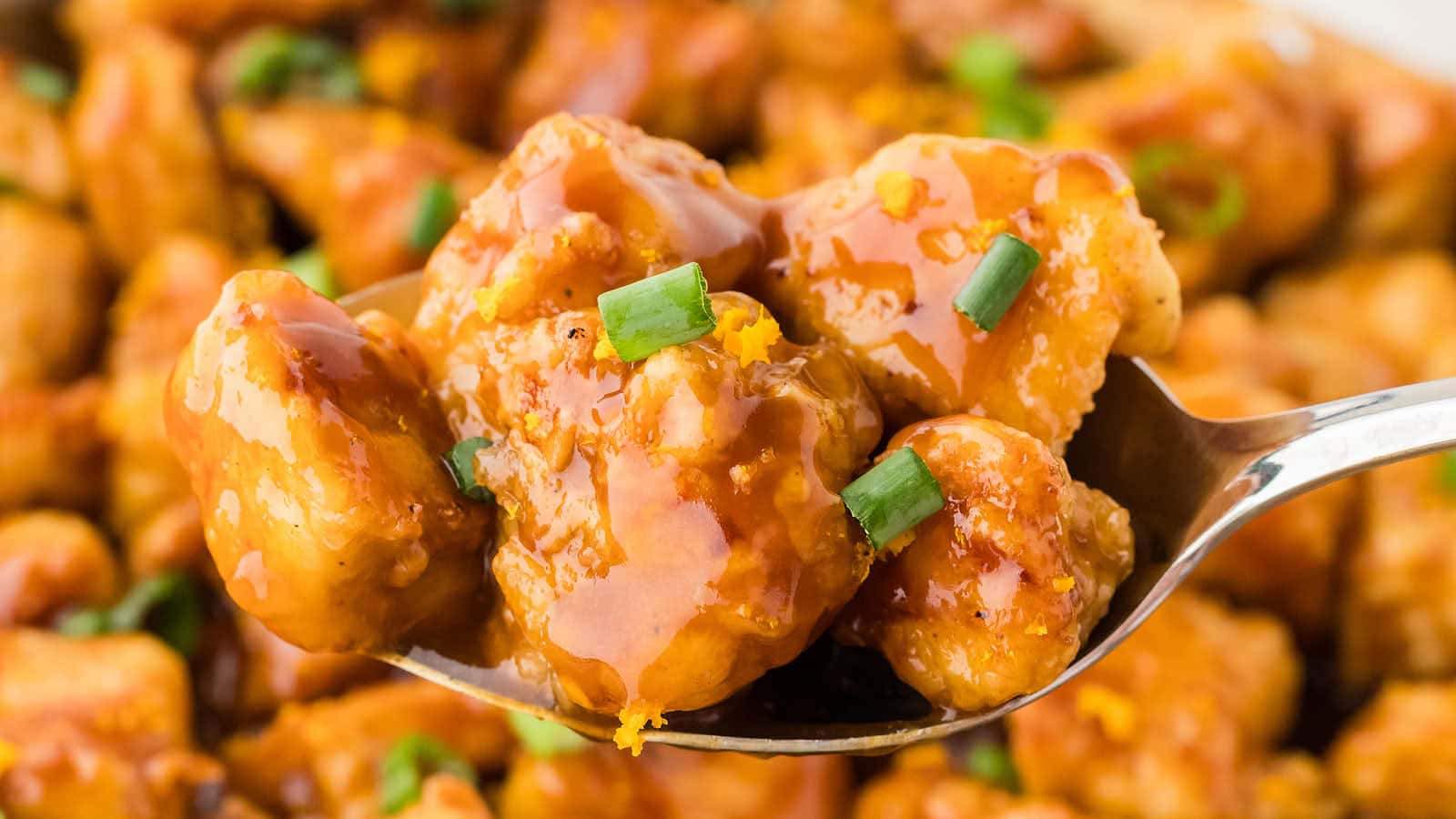 A close-up of a spoon holding pieces of orange chicken topped with sauce and chopped green onions, with more chicken in the background.