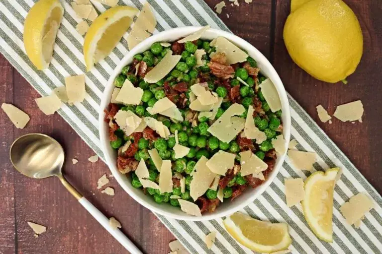 A white bowl filled with green peas, bacon pieces, and shaved parmesan cheese, placed on a striped cloth with lemon wedges and a spoon nearby.
