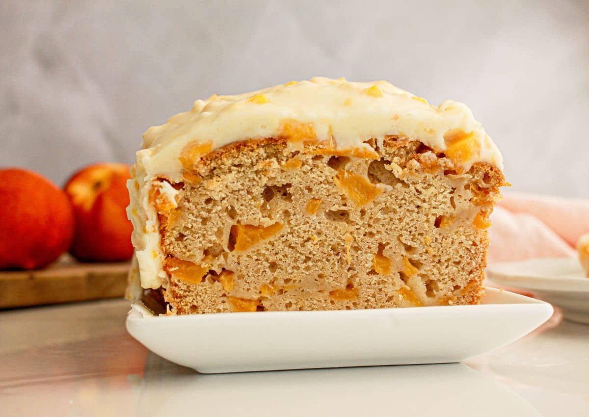 A slice of peach bread with chunks of peach and frosting on top, served on a white plate, with whole peaches in the background.