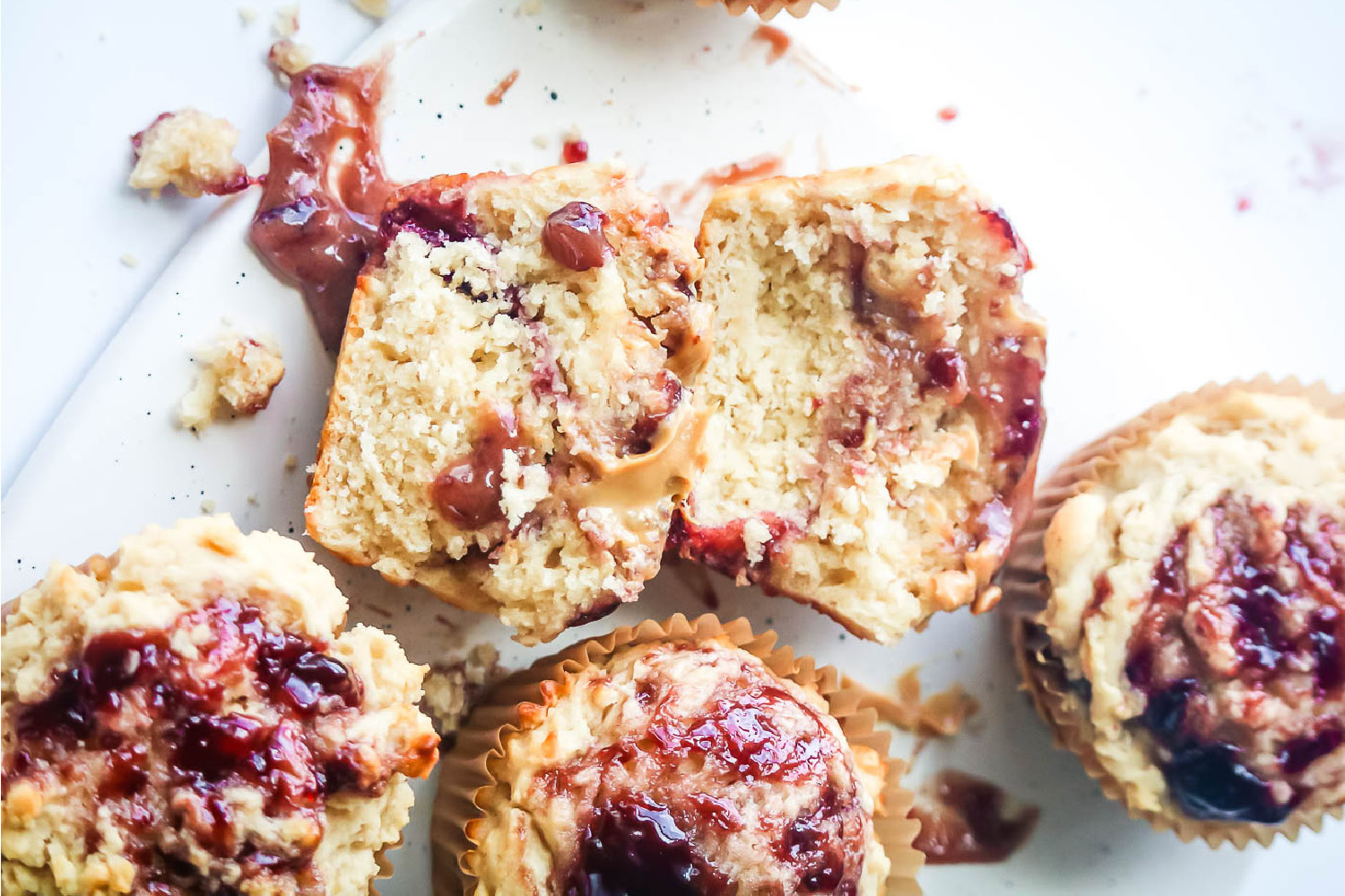 Several baked muffins with peanut butter and jelly swirled on top sit on a white surface, one muffin is cut in half to show the inside texture.
