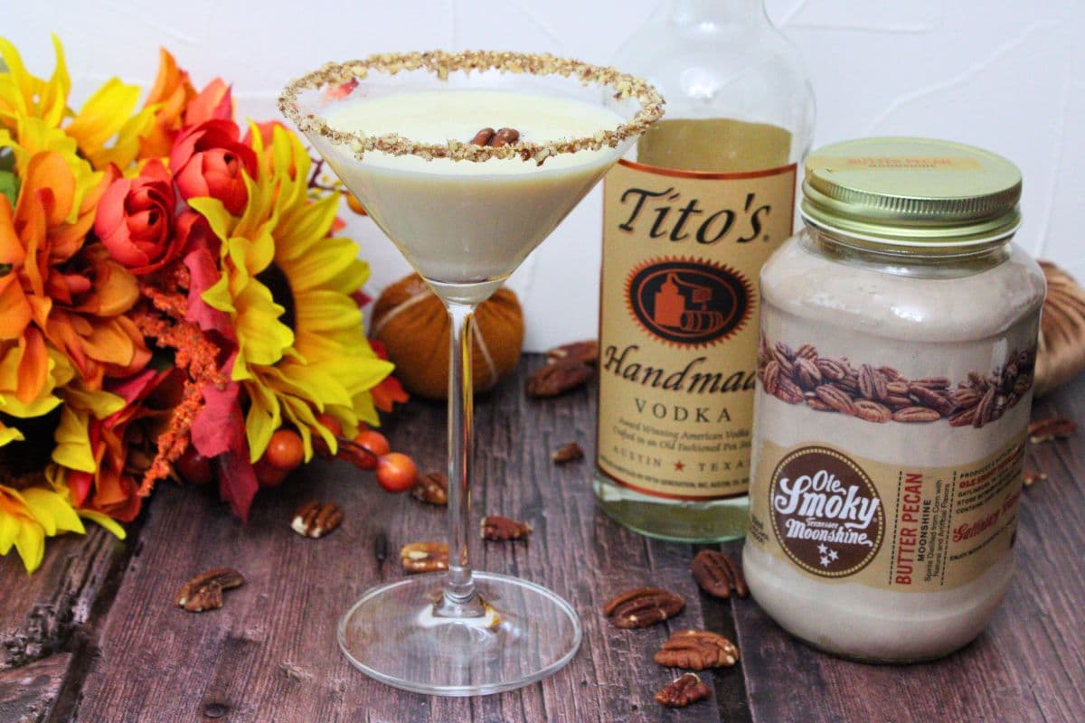 A martini glass with a nut-crusted rim holds a creamy drink, placed next to Titoโs vodka, Ole Smoky Butter Pecan Moonshine, pecans, and autumn-themed decorations.