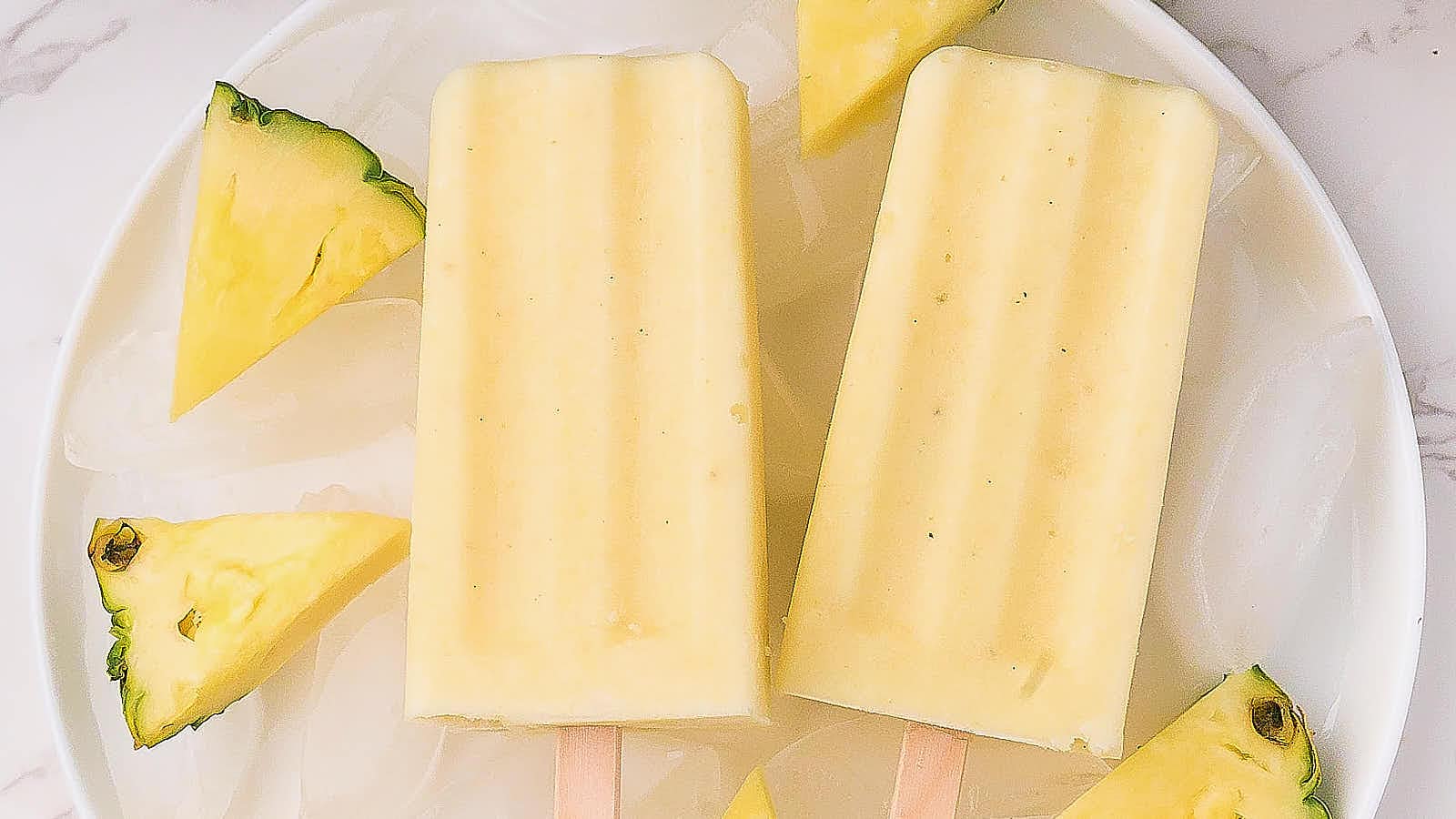 Two yellow popsicles on a white plate with ice cubes, surrounded by pineapple wedges.