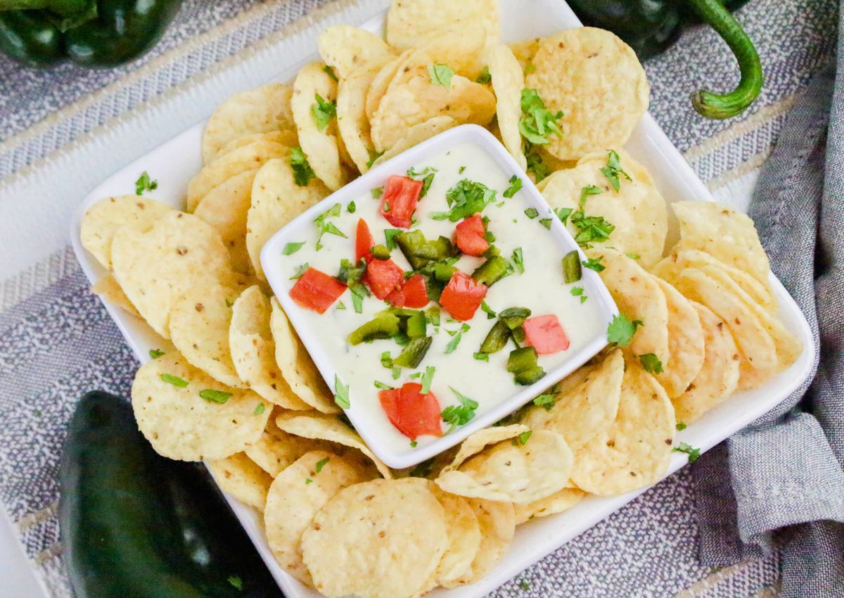 A square white bowl of queso dip topped with chopped tomatoes and peppers is surrounded by round tortilla chips on a white tray.