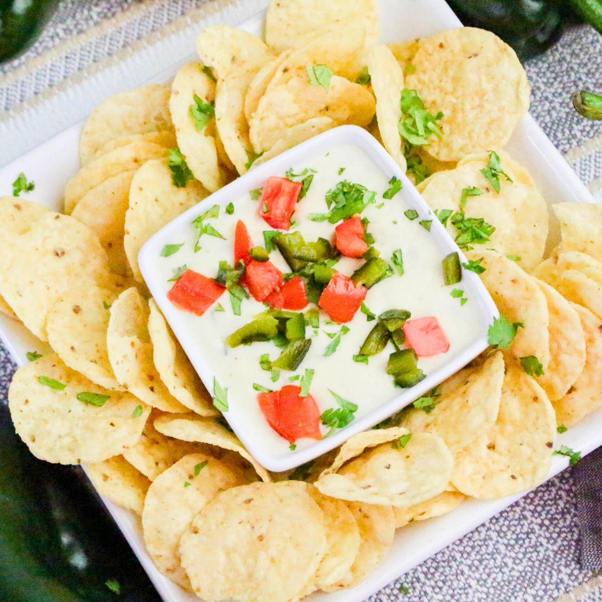 A square bowl of white dip topped with chopped tomatoes and jalapeños is surrounded by round tortilla chips, garnished with cilantro.