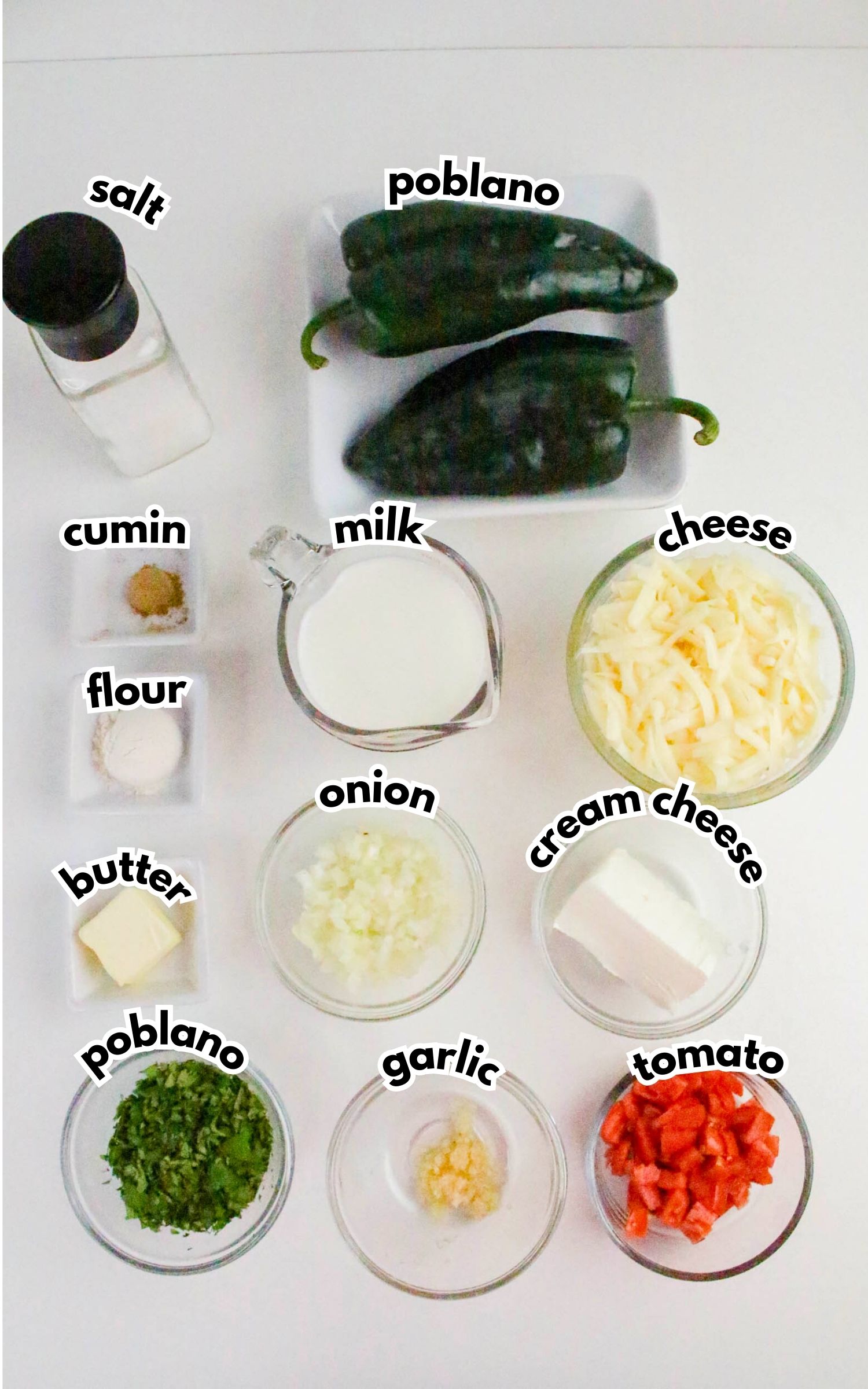 Assorted ingredients on a white surface: poblano peppers, salt, cumin, flour, butter, chopped onion, milk, shredded cheese, cream cheese, chopped poblano, minced garlic, and diced tomato.