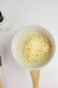 A white saucepan with grated cheese on top of a creamy mixture sits on a white countertop next to a salt shaker.