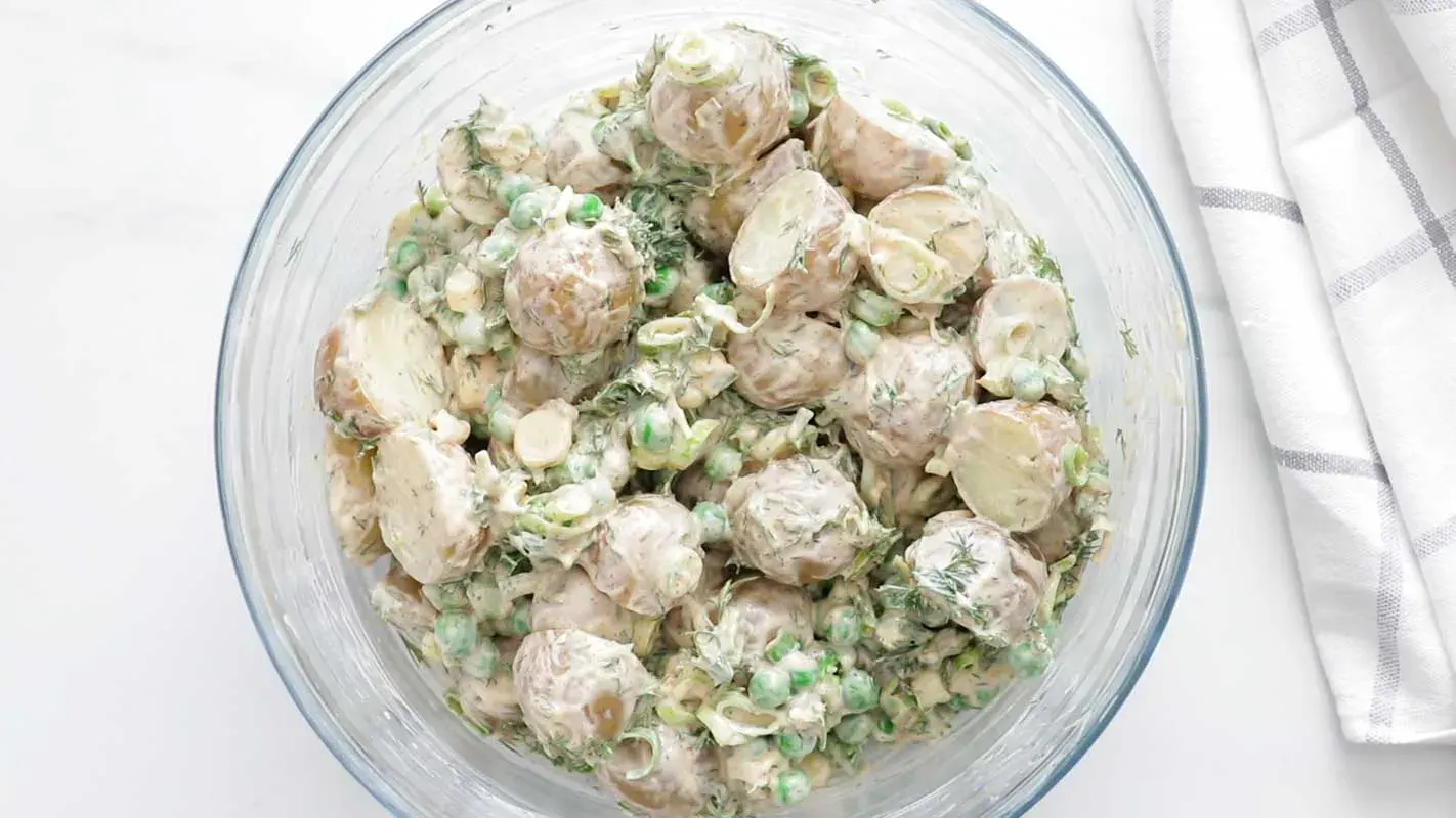 A clear bowl filled with creamy potato salad containing halved potatoes, green peas, chopped herbs, and sliced scallions on a white surface.