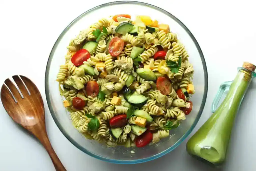 A glass bowl filled with rotini pasta salad, cherry tomatoes, cucumber, cheese cubes, and herbs, with a wooden utensil and a bottle of green dressing beside it.