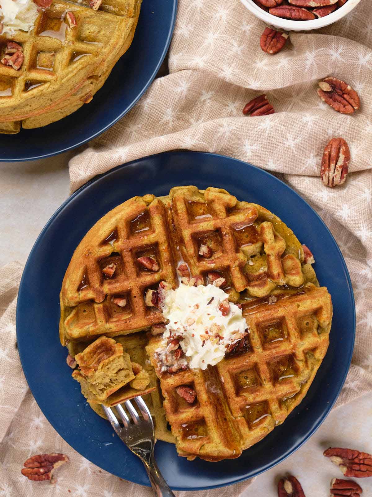A plate with two sliced waffles topped with whipped cream and chopped pecans, surrounded by scattered pecans on a beige cloth.