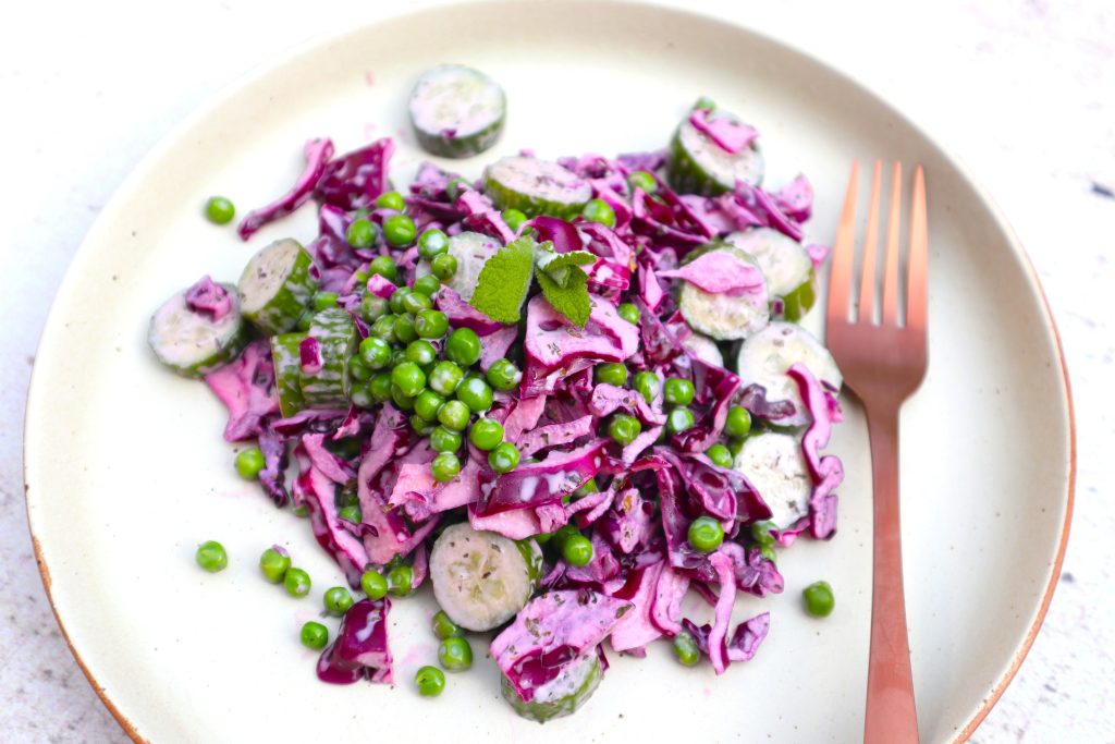 A plate of salad with sliced cucumber, shredded red cabbage, green peas, and a creamy dressing, garnished with fresh herbs. A copper fork is placed on the side.