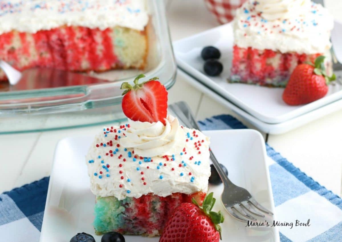 A square slice of red, white, and blue poke cake topped with whipped cream, sprinkles, and a halved strawberry, served on a white plate with fresh berries. The rest of the cake is in a glass dish.