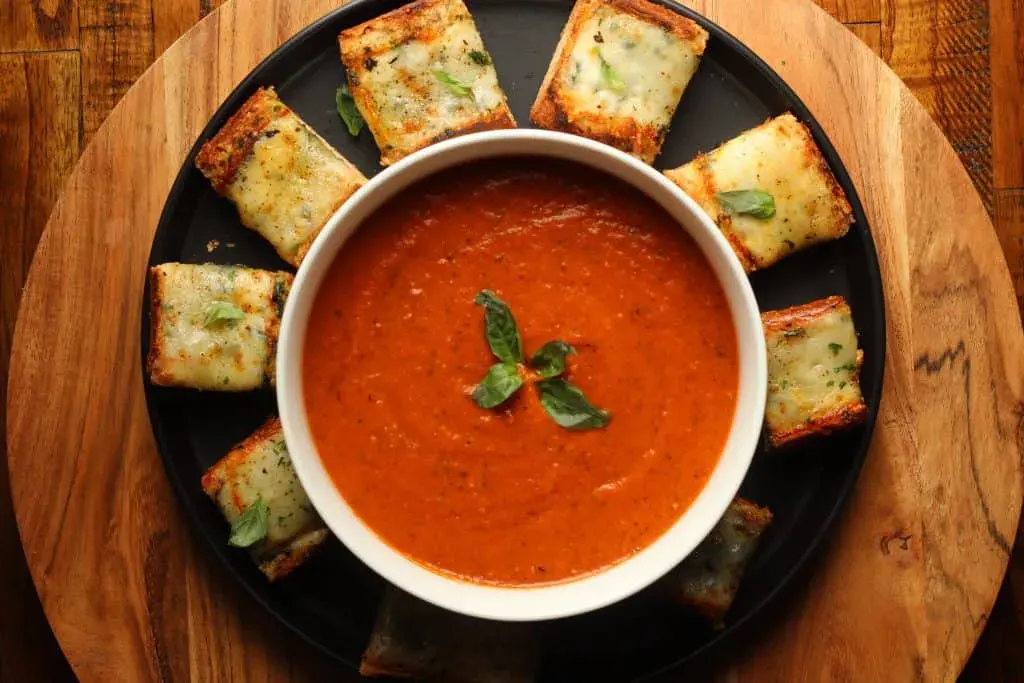 A bowl of tomato soup garnished with basil is surrounded by rectangular pieces of cheesy garlic bread on a round black plate.