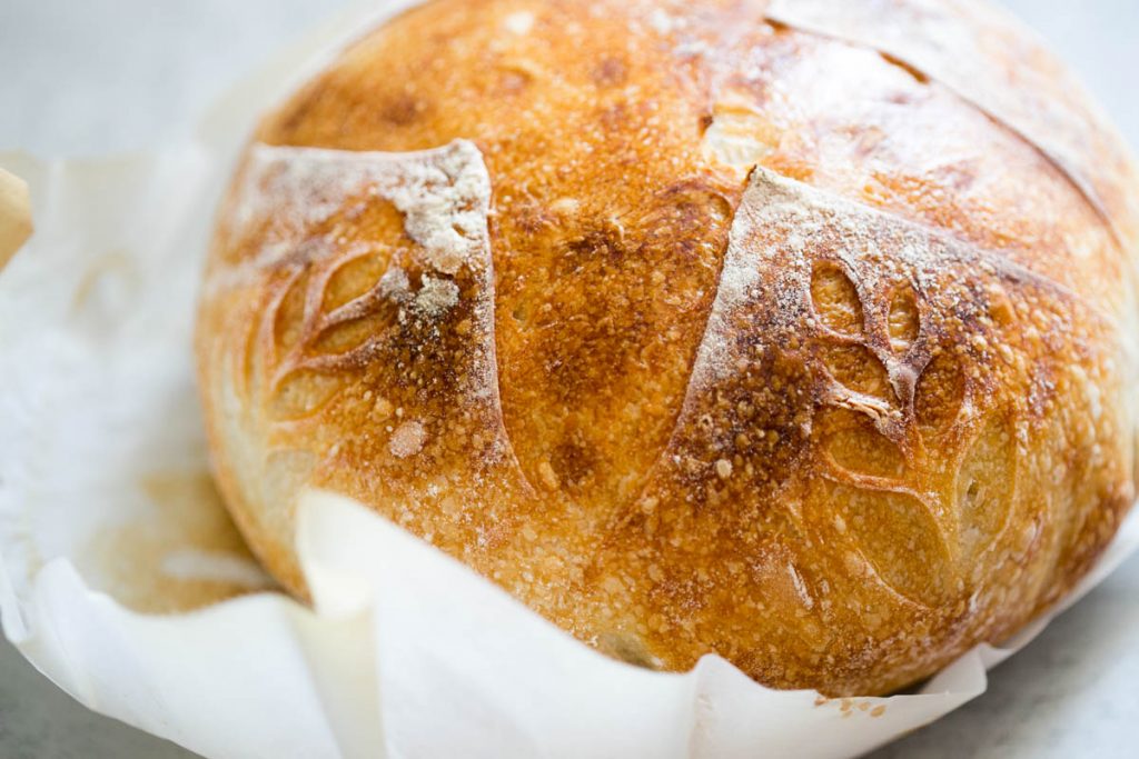 A round loaf of artisan sourdough bread with a golden crust and leaf-shaped scoring, resting on parchment paper.