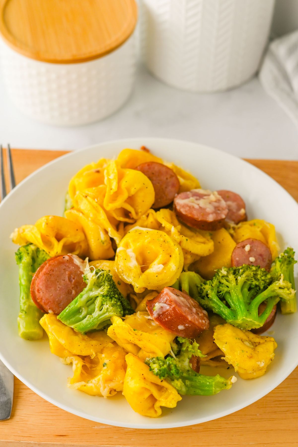 A white plate with cheese tortellini, sliced sausage, and broccoli on a wooden surface, with white containers in the background.