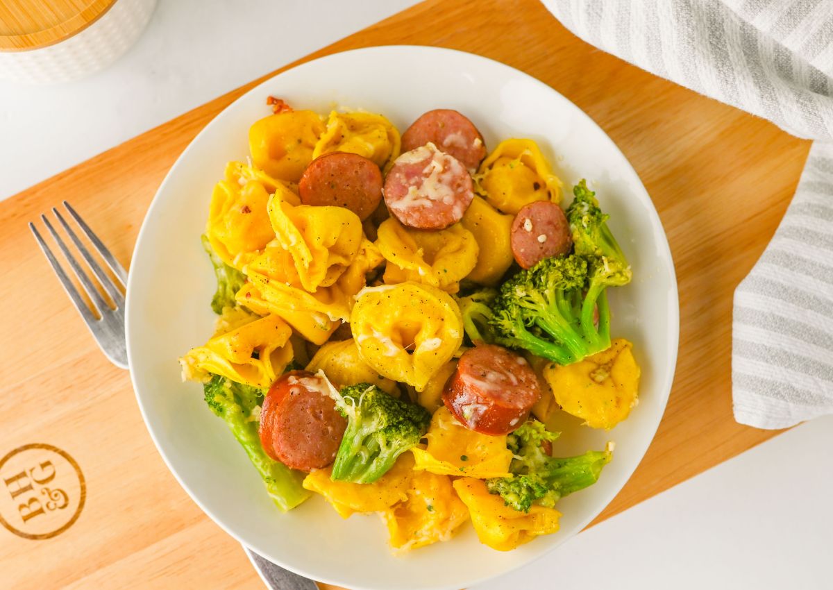 A plate of tortellini pasta with broccoli florets and sliced sausage, topped with grated cheese, on a wooden board next to a fork and striped cloth.