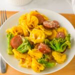 A white plate with cheese tortellini, sliced sausage, and broccoli, placed on a wooden surface beside a fork.