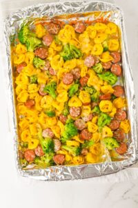 A foil-lined baking sheet filled with cooked cheese tortellini, sliced sausage, and broccoli florets.