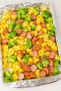 A foil-lined baking sheet with uncooked tortellini pasta, sliced sausage, and broccoli florets arranged evenly.
