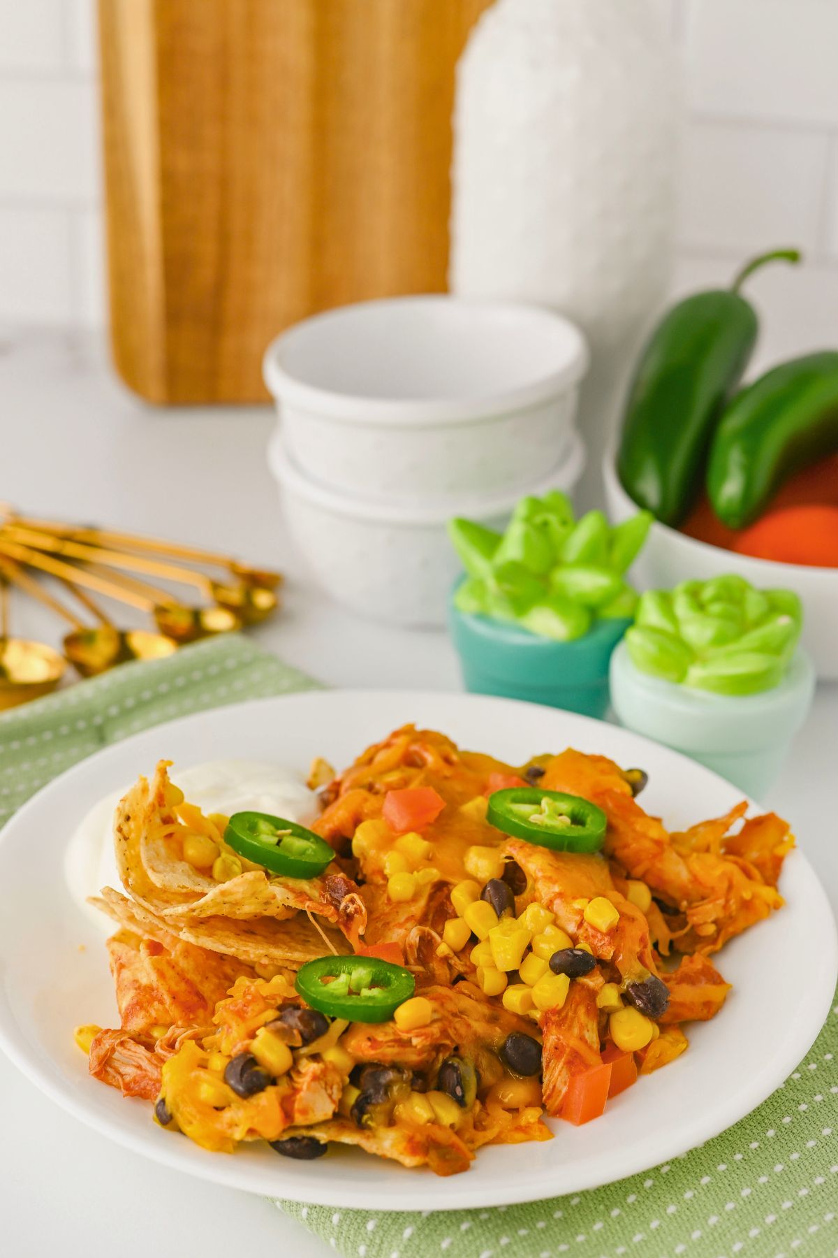 A plate of chicken nachos topped with melted cheese, corn, black beans, diced tomatoes, and sliced jalapeños sits on a table with bowls, gold utensils, and vegetables in the background.