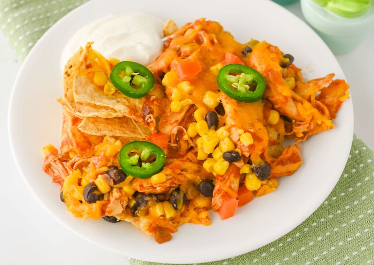 A plate of nachos topped with melted cheese, corn, black beans, diced tomatoes, jalapeño slices, shredded chicken, and a side of sour cream.
