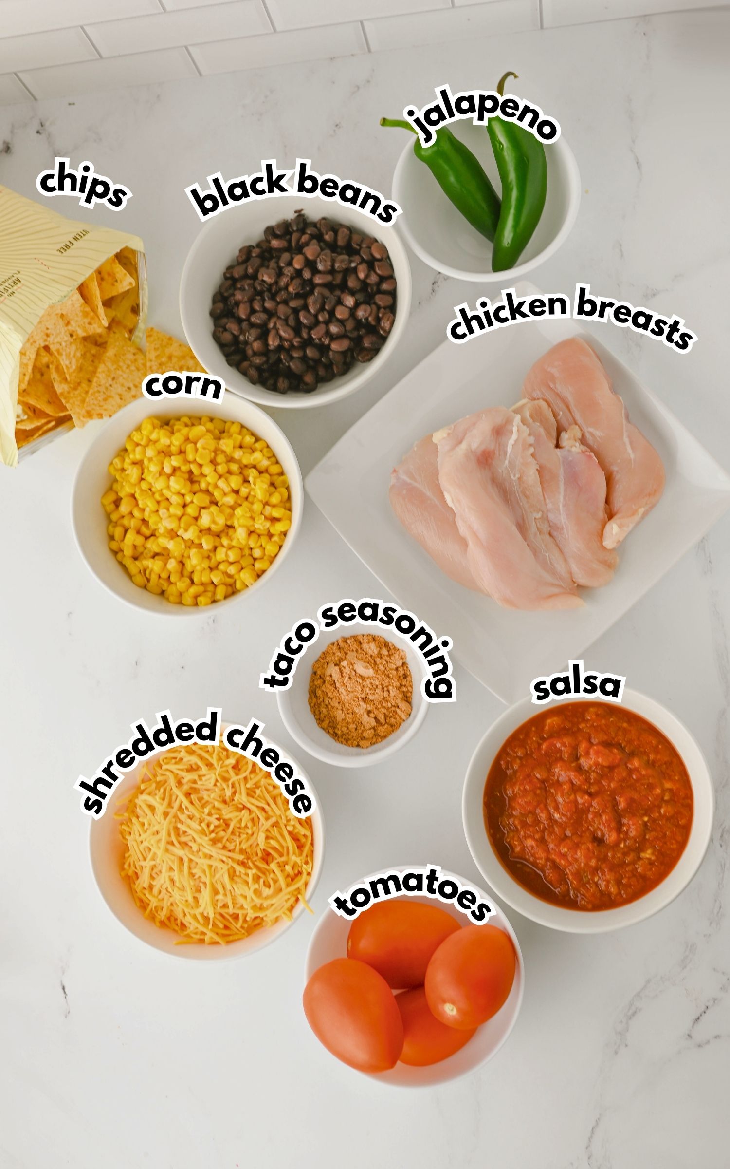 Bowls containing chips, black beans, corn, shredded cheese, taco seasoning, salsa, fresh tomatoes, jalapenos, and raw chicken breasts arranged on a white countertop.