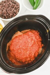 A slow cooker filled with a layer of ground meat and topped with red salsa, surrounded by bowls of black beans, corn, and jalapeños.