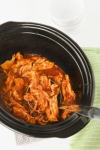 Shredded chicken in red sauce inside a black slow cooker, with a metal serving spoon and a green cloth nearby.
