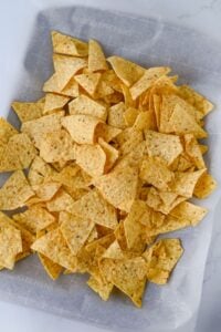 A pile of yellow corn tortilla chips scattered on a sheet of parchment paper.