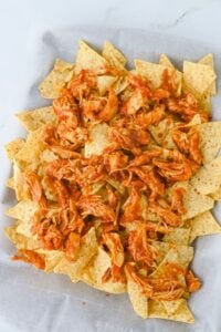 Tortilla chips topped with shredded barbecue chicken, arranged on a sheet of parchment paper.