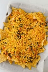 A baking sheet lined with parchment paper holds tortilla chips topped with shredded cheddar cheese, black beans, and corn.
