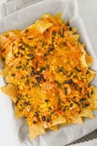 A tray of nachos topped with melted cheese, black beans, corn, and shredded chicken on a piece of parchment paper.